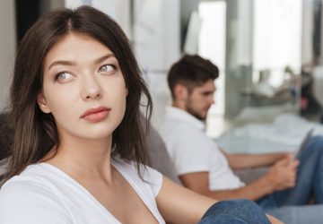 Sad thoughtful young woman sitting while her boyfriend using tablet